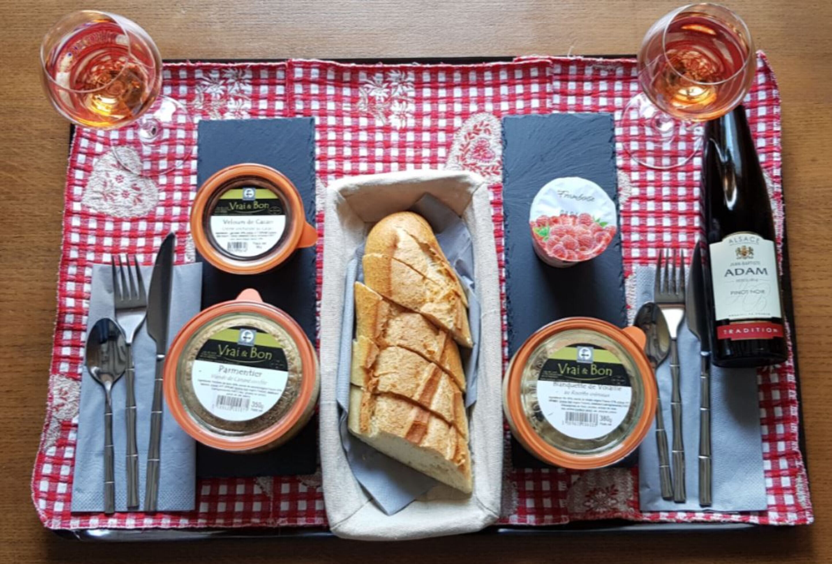 Uma mesa posta com uma baguete, dois potes de geleia Vi&Bon, uma garrafa de vinho Adam e uma toalha de mesa xadrez.