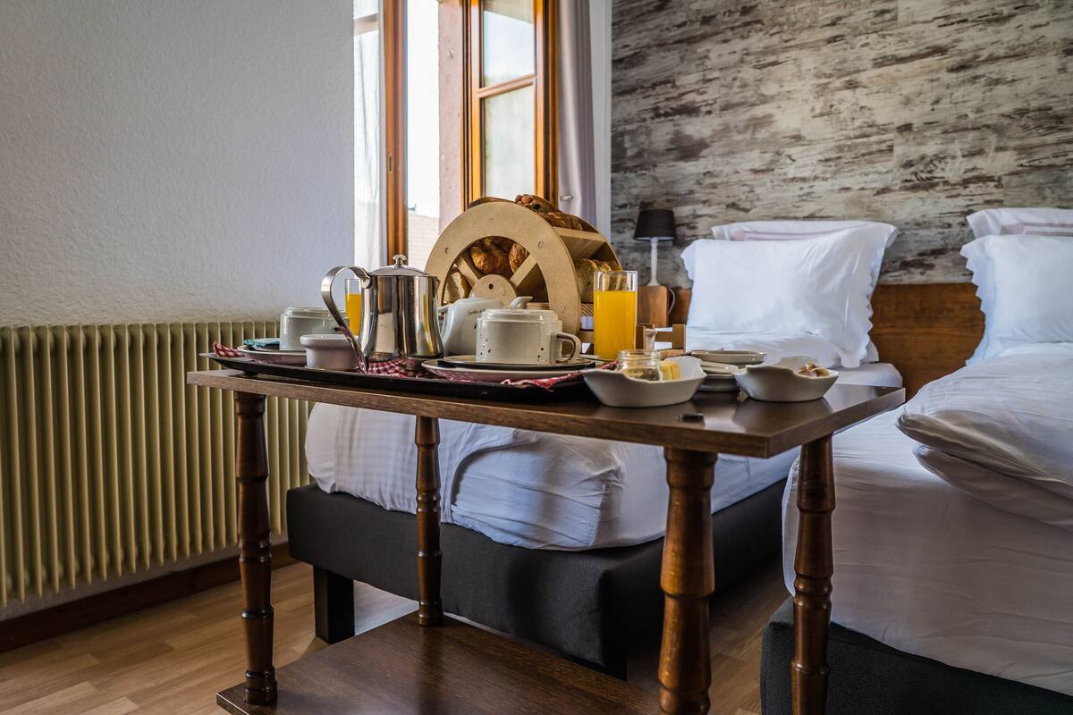 Una mesa de desayuno ordenada con una tetera, tazas y un frutero en una habitación con una cama y una pared rústica.