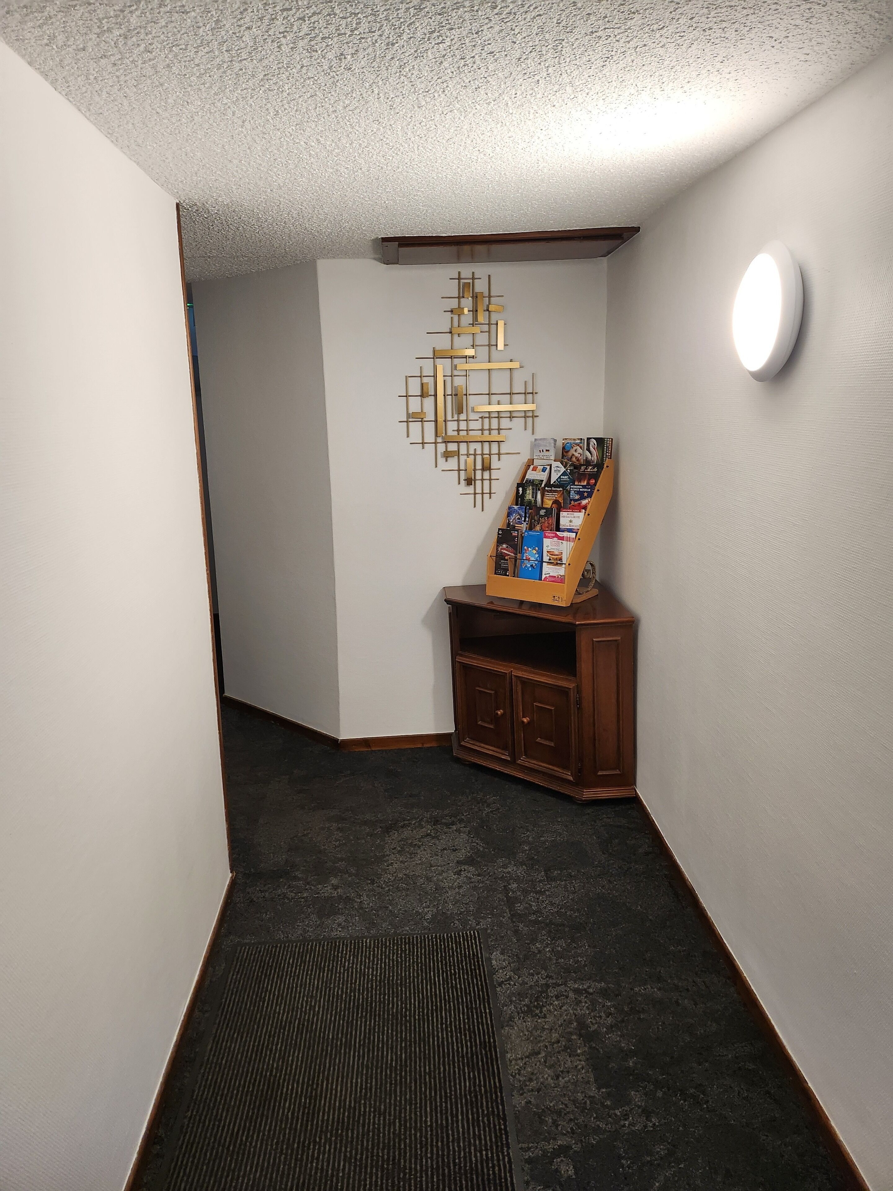 A corner room with a dark carpet, a wooden cabinet with magazines, a wall-mounted light fixture, and a geometric wall decoration.