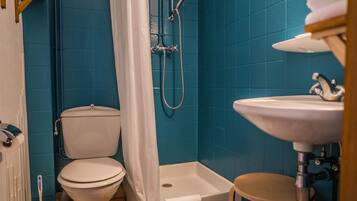 A bathroom with a toilet, shower, sink, and a small stool.