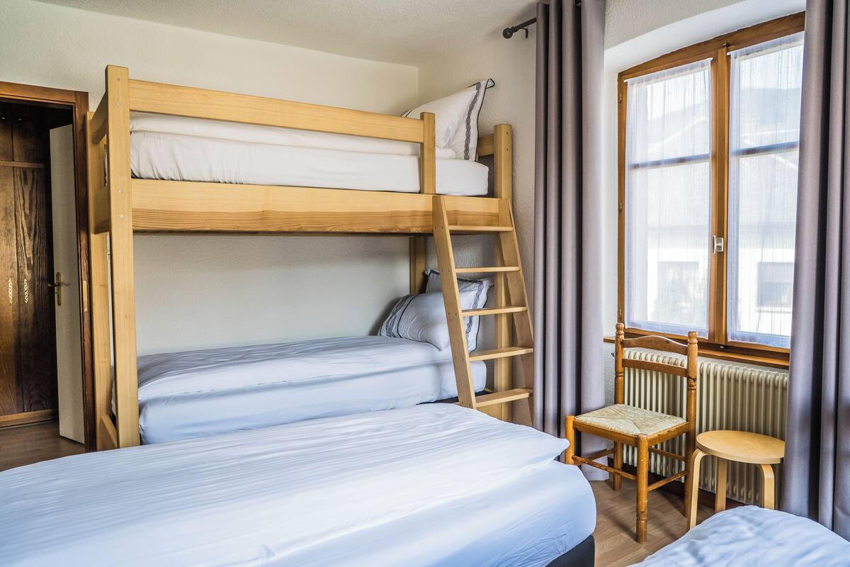 Habitación con literas de madera, una escalera, una silla y una ventana con cortinas.