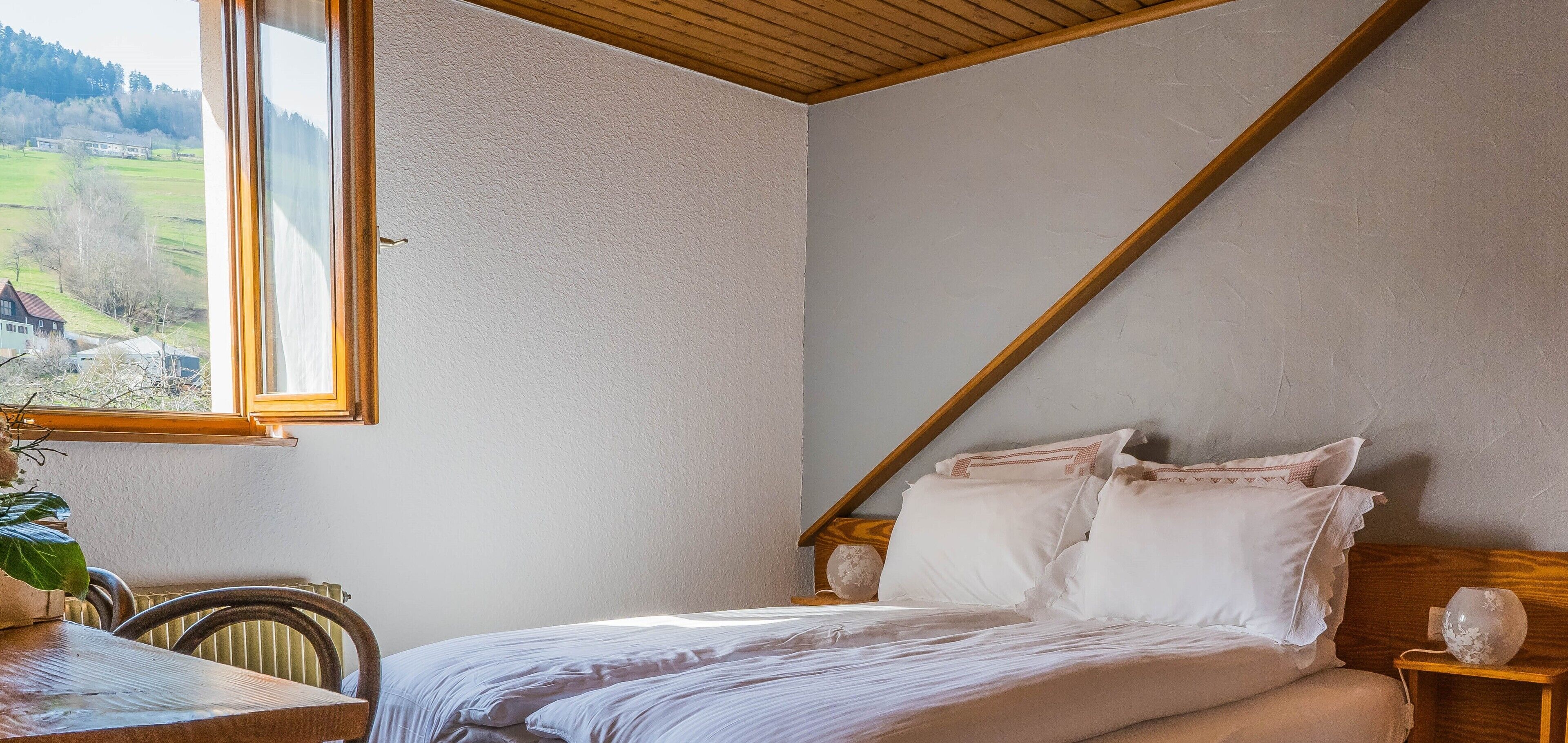 A bedroom with a bed, wooden headboard, bedside tables, and a window with a view of a house and greenery.