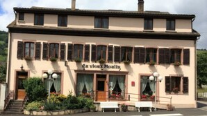 A two-story building with a sign reading "La Vieux Moulin." It features a balcony with potted plants, a front entrance with steps, and a bench outside. - Au Vieux Moulin (Lapoutroie)