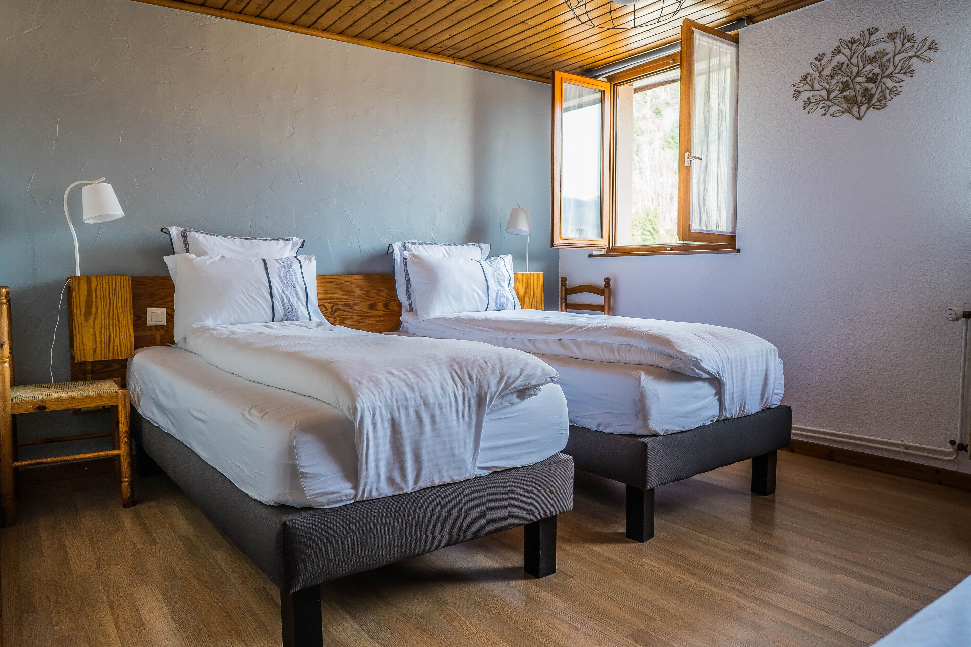 Una habitación con dos camas, suelo de madera, una ventana con cortinas y una lámpara fijada a la pared.