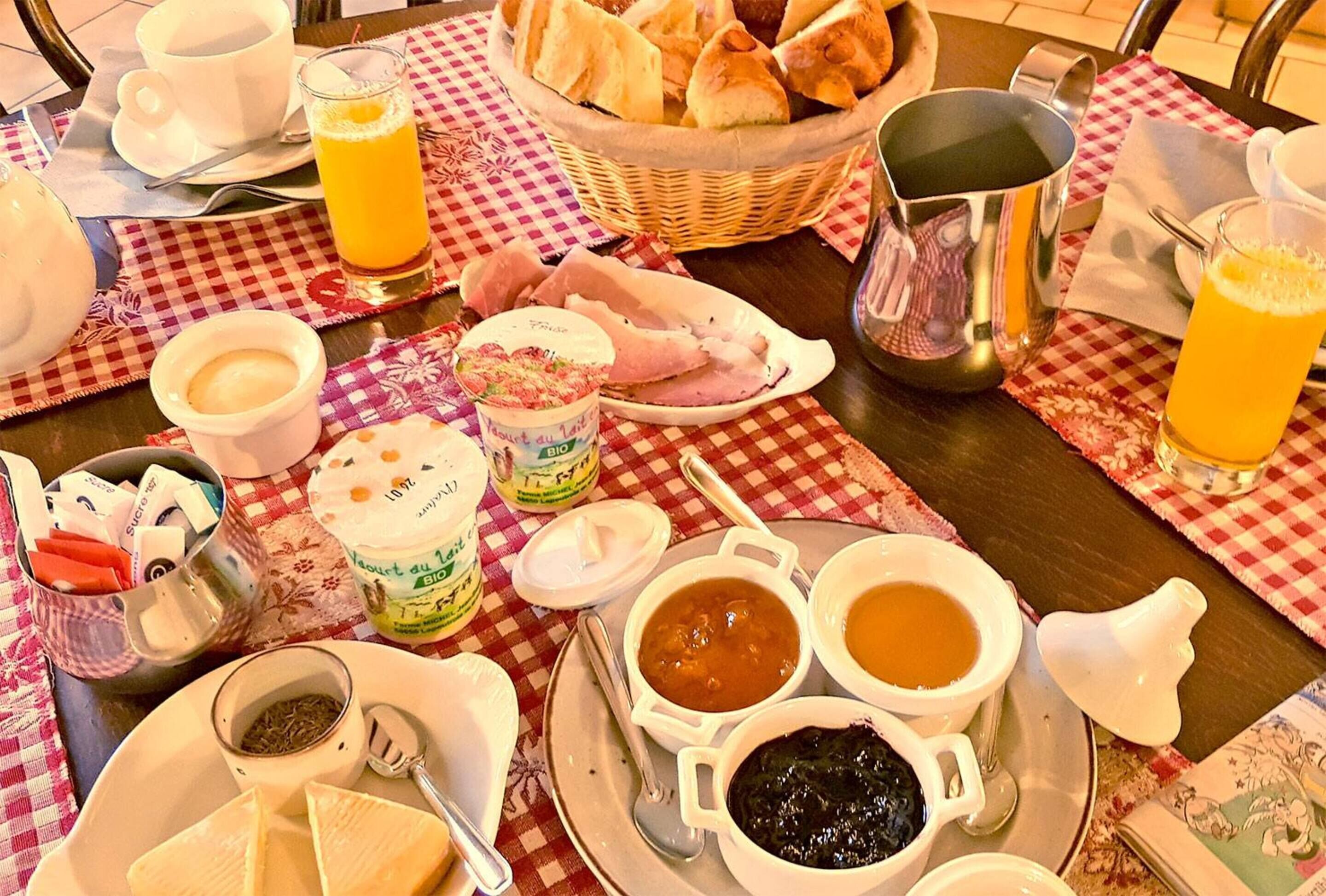 Aamiaispöytä, jonka päällä on ruutukuosinen pöytäliina, ja jolla on tarjolla erilaisia aamiaisruokia, kuten leipää, leivonnaisia ja juomia, sekä kori täynnä croissantteja.