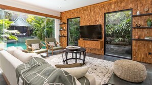 A modern living room with a sofa, chairs, a TV, and a pool view. - Villa Suvitha by Villa Finder (Seminyak)