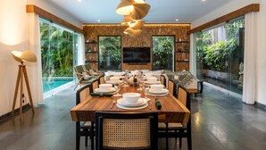 A dining area with a wooden table set for a meal, surrounded by chairs, a large TV, and a view of a swimming pool. - Villa Suvitha by Villa Finder (Seminyak)