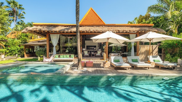A poolside area with lounge chairs, umbrellas, and a thatched-roof structure. - Villa Desa Roro Dua by Villa Finder (Canggu)