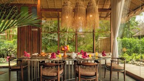 A dining area with a table set for four, surrounded by wooden chairs, with a tropical backdrop and a hanging light fixture above. - Villa Desa Roro Dua by Villa Finder (Canggu)