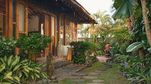 A traditional wooden house with a thatched roof, surrounded by lush tropical vegetation and a stone pathway. - Villa Desa Roro Dua by Villa Finder (Canggu)