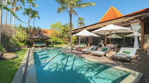 A poolside area with lounge chairs and umbrellas, surrounded by palm trees and a thatched-roof building. - Villa Desa Roro Dua by Villa Finder (Canggu)