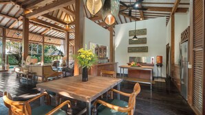 A spacious dining area with a wooden table and chairs, a vase of yellow flowers, and a kitchen in the background. - Villa Desa Roro Dua by Villa Finder (Canggu)