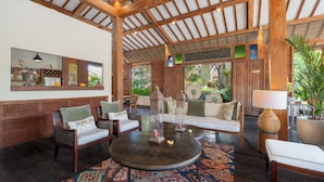A spacious lounge area with a round wooden table, cushioned seating, and a decorative mirror. - Villa Desa Roro Dua by Villa Finder (Canggu)