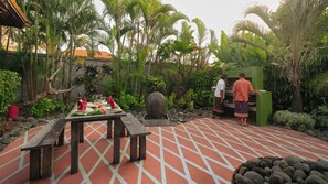 A dining area with a table set for two, surrounded by tropical plants and a stone fountain. - Villa Desa Roro Dua by Villa Finder (Canggu)