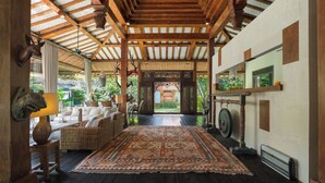 A traditional wooden interior with a large rug, wicker furniture, and a view to an outdoor garden. - Villa Desa Roro by Villa Finder (Canggu)