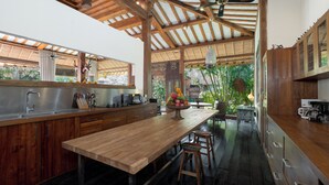 A modern kitchen with wooden cabinets, a large island, and a view of lush greenery outside. - Villa Desa Roro by Villa Finder (Canggu)