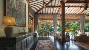 A spacious room with a high ceiling, wooden beams, a large painting, a dining area with a table and chairs, and a view of lush greenery outside. - Villa Desa Roro by Villa Finder (Canggu)