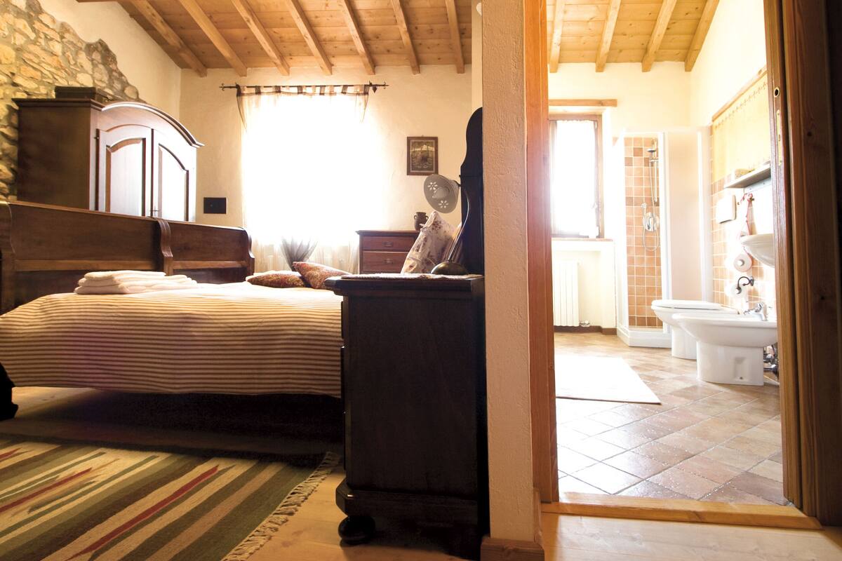 A bedroom with a wooden bed, a stone wall, a wooden dresser, and a bathroom visible through an open door.
