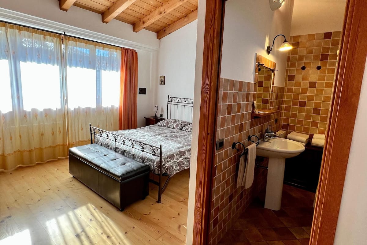 A bedroom with a bed, a bench, and a bathroom with a sink and mirror.