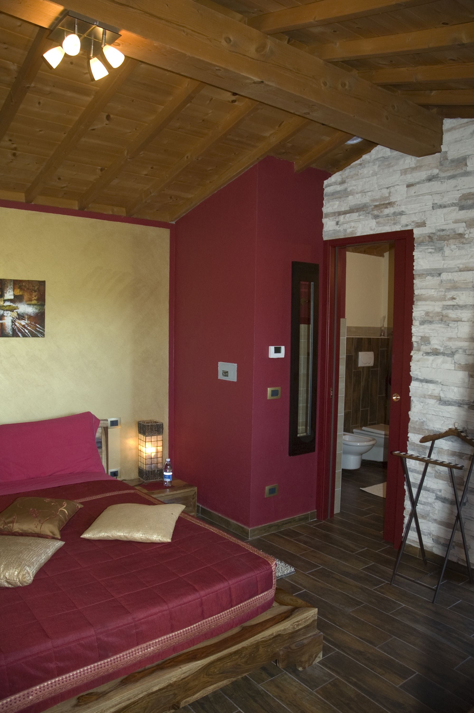 A bedroom with a wooden ceiling, a red wall, a bed with a red and beige bedspread, a nightstand with a lamp, and a bathroom visible through an open door.