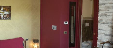A bedroom with a wooden ceiling, a red wall, a bed with a red and beige bedspread, a nightstand with a lamp, and a bathroom visible through an open door.