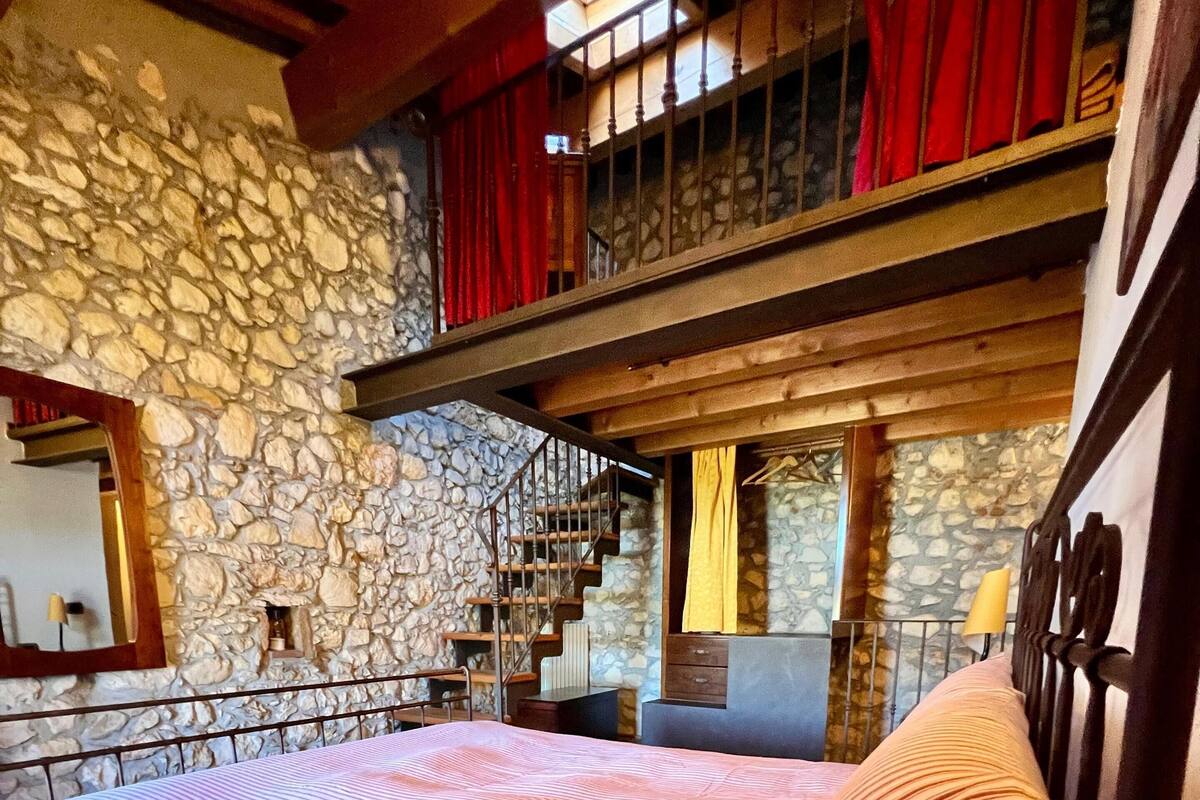 A bedroom with a stone wall, a bed with striped bedding, a wooden headboard, and a balcony with red curtains.