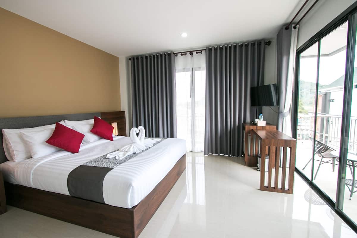 A modern hotel room with a large bed, a wooden side table, a TV, and a balcony with outdoor seating.