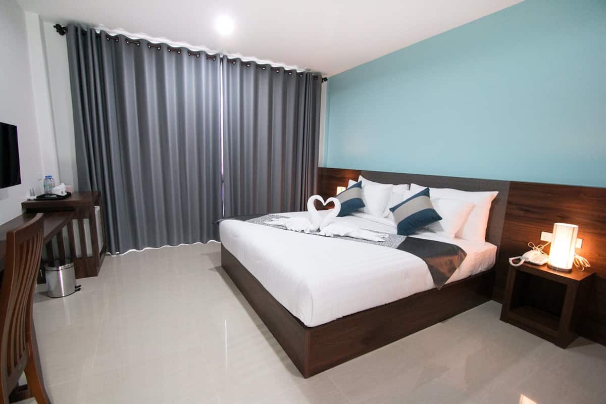 A hotel room with a large bed, a wooden nightstand with a lamp, a desk with a chair, and a TV.