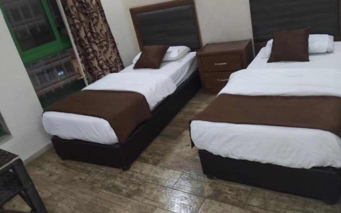 Une chambre d’hôtel avec deux lits, chacun doté d’une tête de lit marron et de draps blancs. Une petite table de chevet en bois se trouve entre les deux lits, et une fenêtre à cadre vert est située sur le côté gauche.