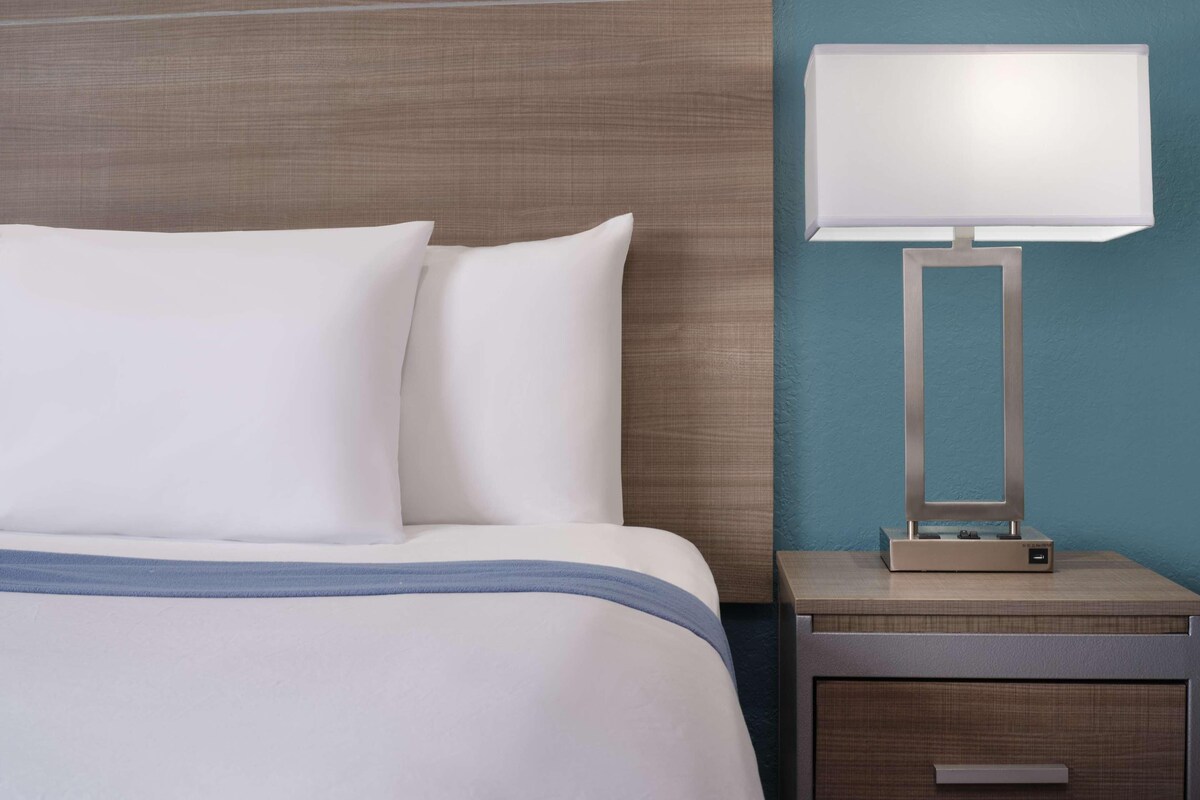 Un lit bien fait, avec du linge de lit blanc et un élément décoratif bleu, une table de chevet avec une lampe, et un mur orné d’un motif texturé.