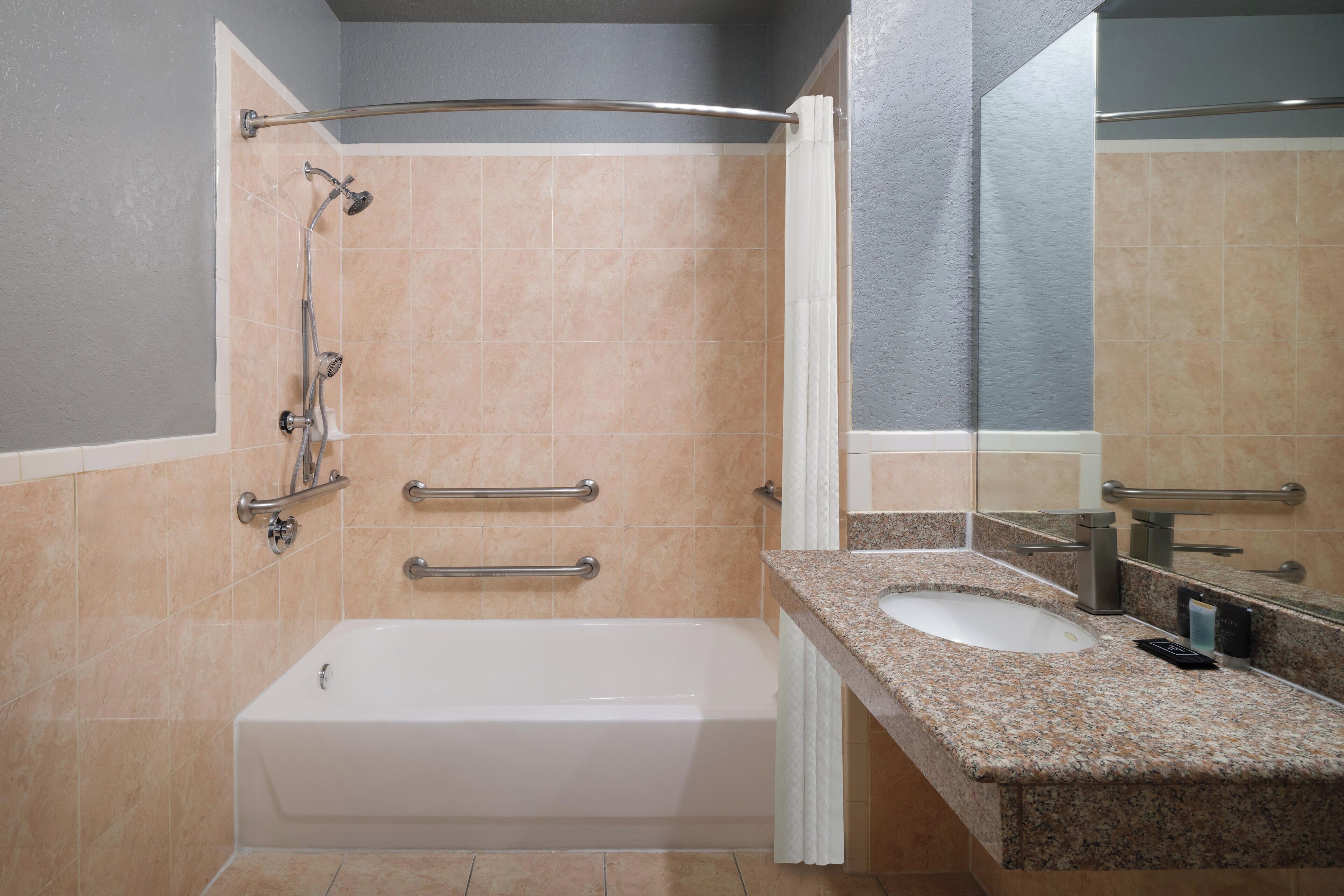 A modern bathroom with a bathtub, a shower with grab bars, a granite countertop sink, and a large mirror.