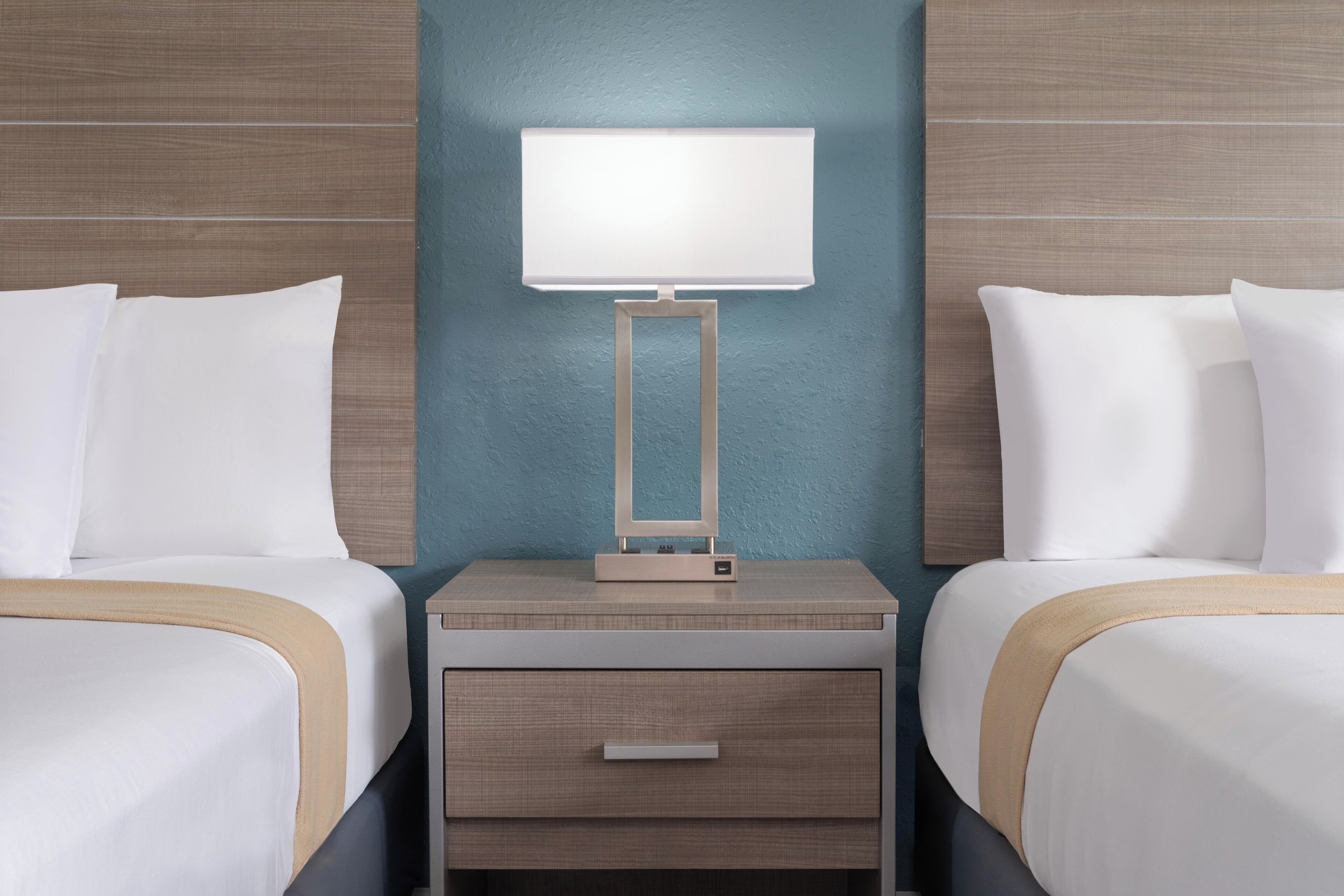 A hotel room with two beds, a bedside table with a lamp, and a drawer.