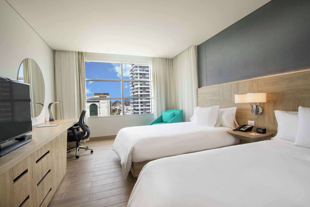 A hotel room with two beds, a desk with a TV, a chair, a nightstand with a lamp, and a view of buildings through the window.