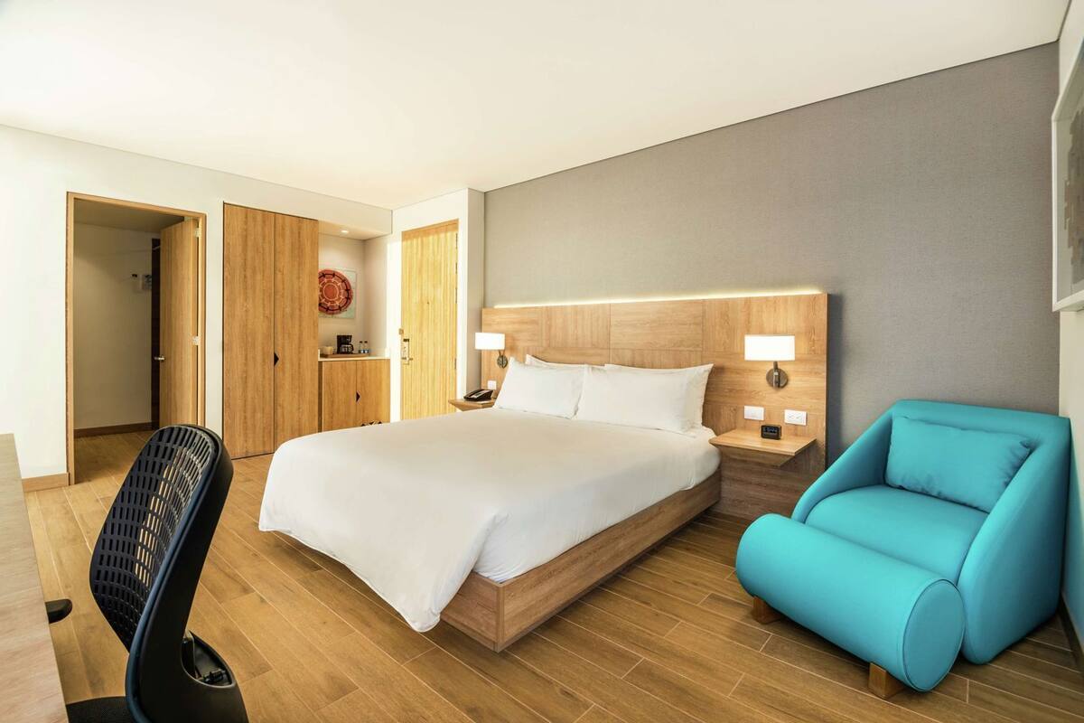 A hotel room with a large bed, a desk, a chair, and a blue armchair.