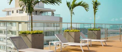 A rooftop lounge with white sun loungers, palm trees, and a view of a modern building.