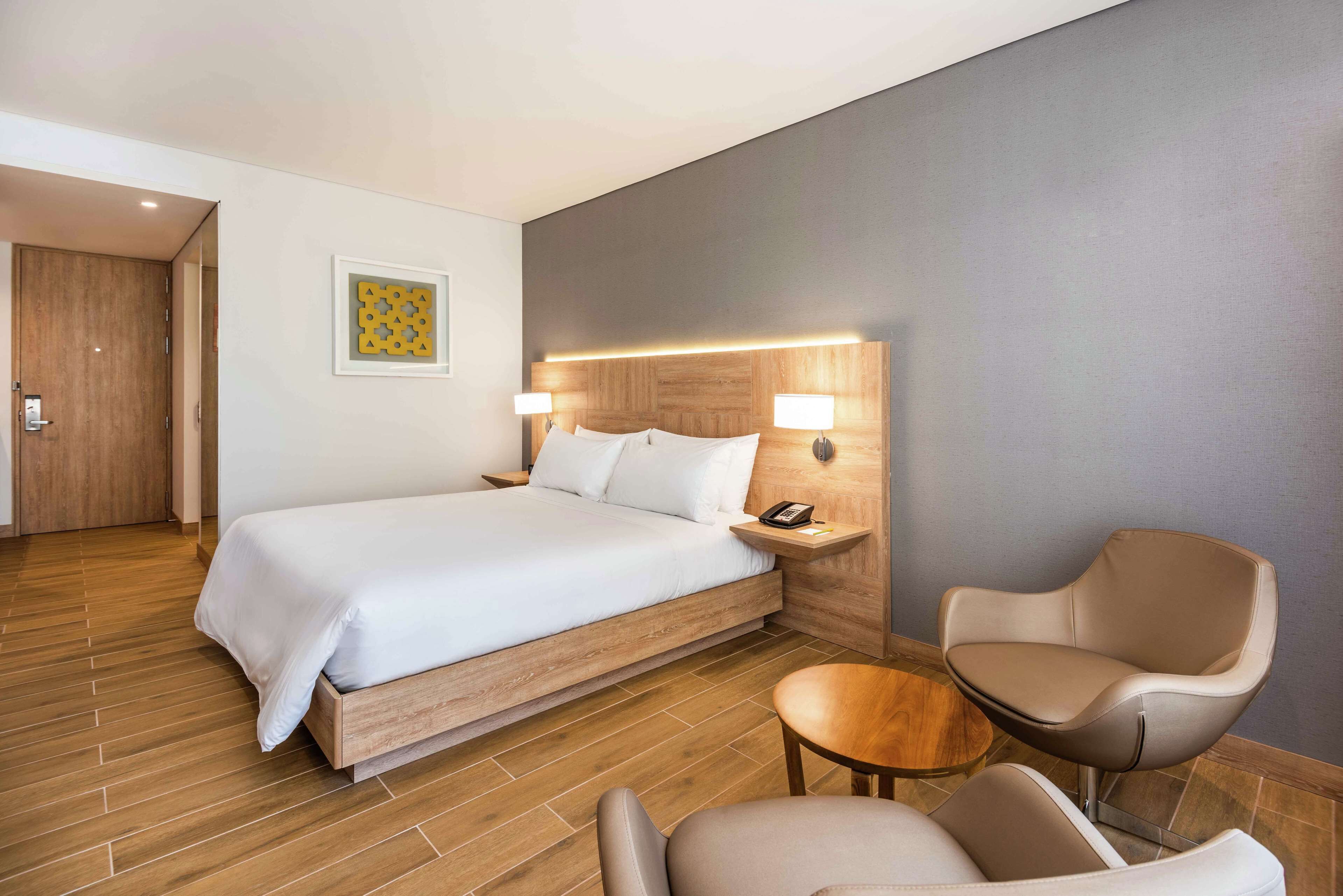 Een moderne hotelkamer met een groot bed, nachtlampjes, een houten hoofdbord, een kleine tafel en een stoel.