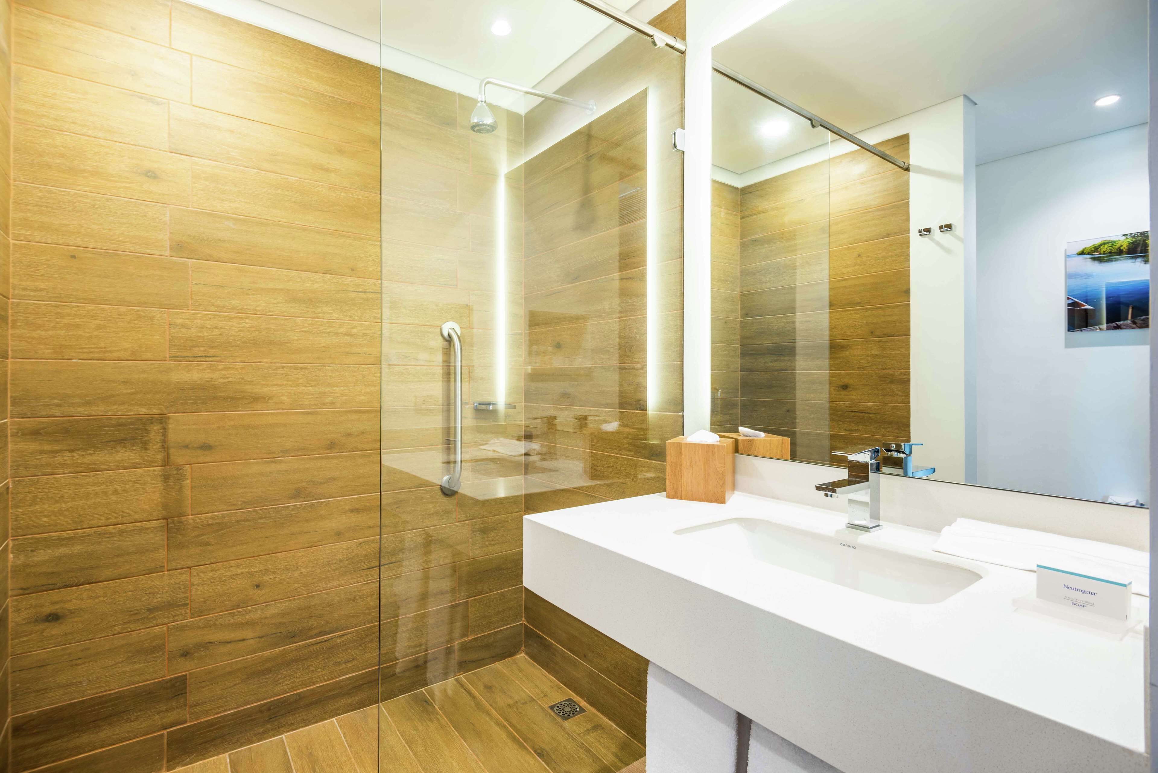 A modern bathroom with a wooden floor, a glass shower enclosure, a white sink, and a large mirror.