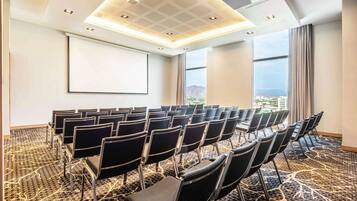 Un salón de conferencias con filas de sillas, una pantalla de proyector y un amplio ventanal con vistas.