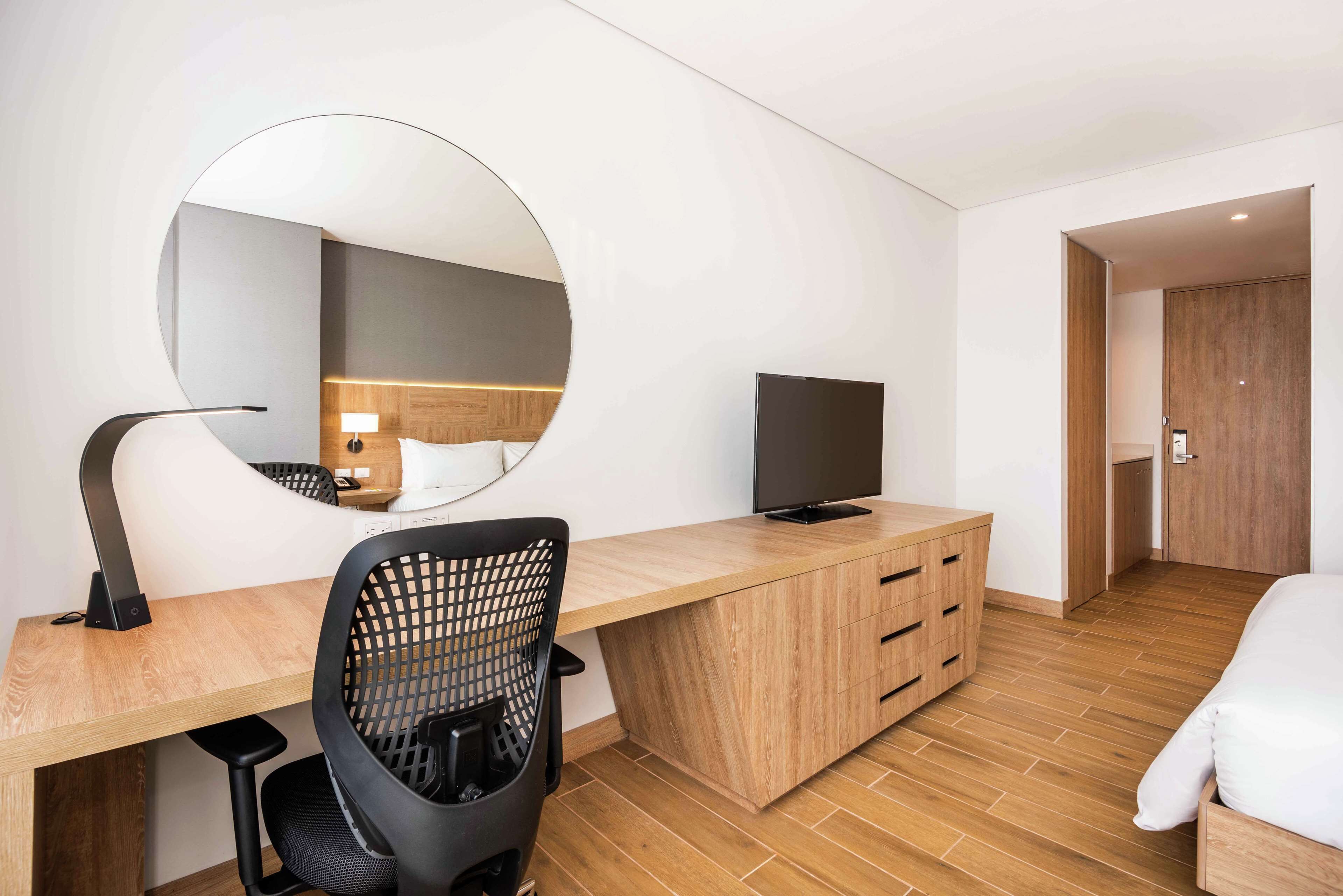 Ett modernt hotellrum med ett skrivbord i trä, en svart kontorsstol, en platt-TV och en stor spegel.