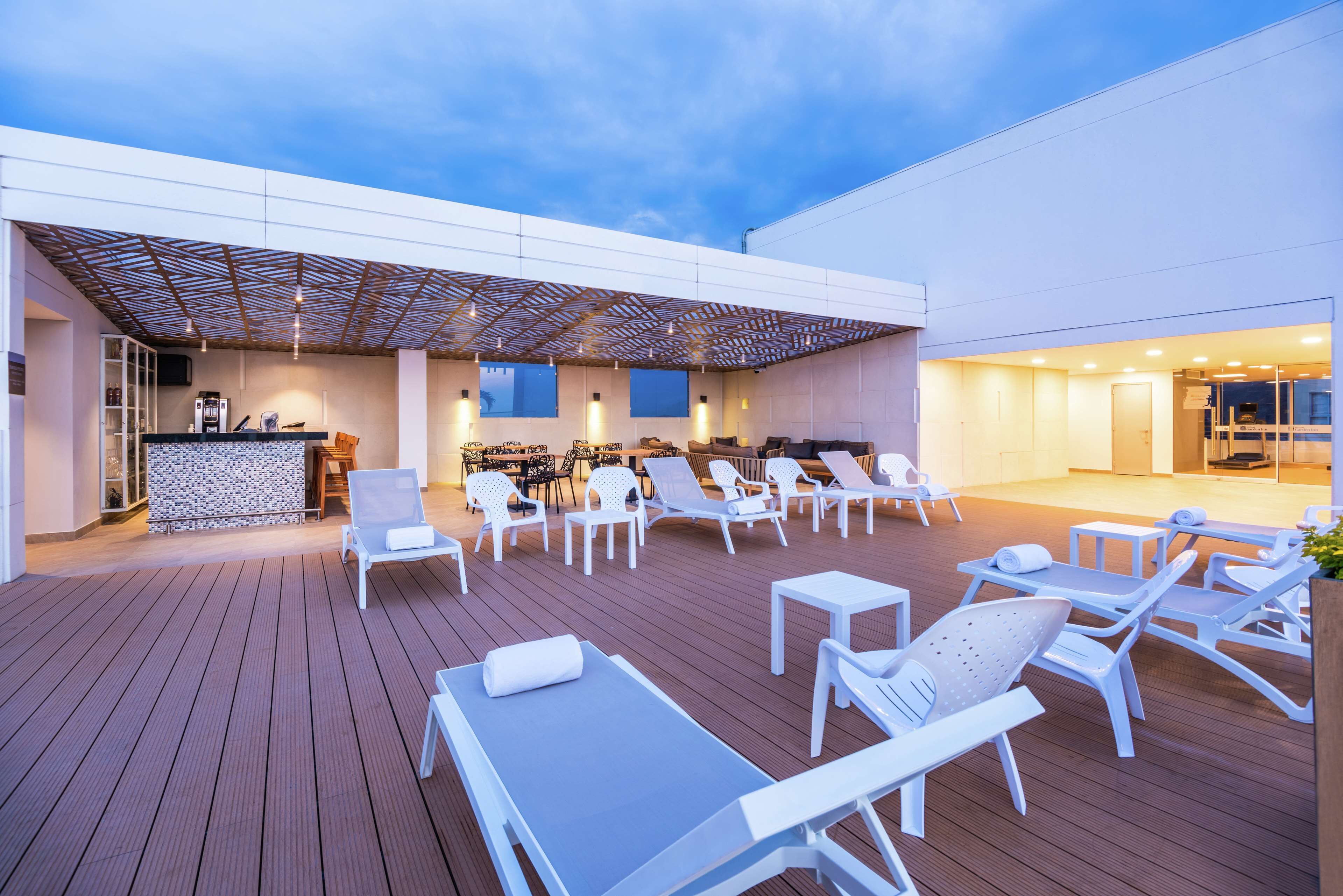 An outdoor deck with white lounge chairs and tables, a bar area, and a covered seating area with a patterned ceiling.