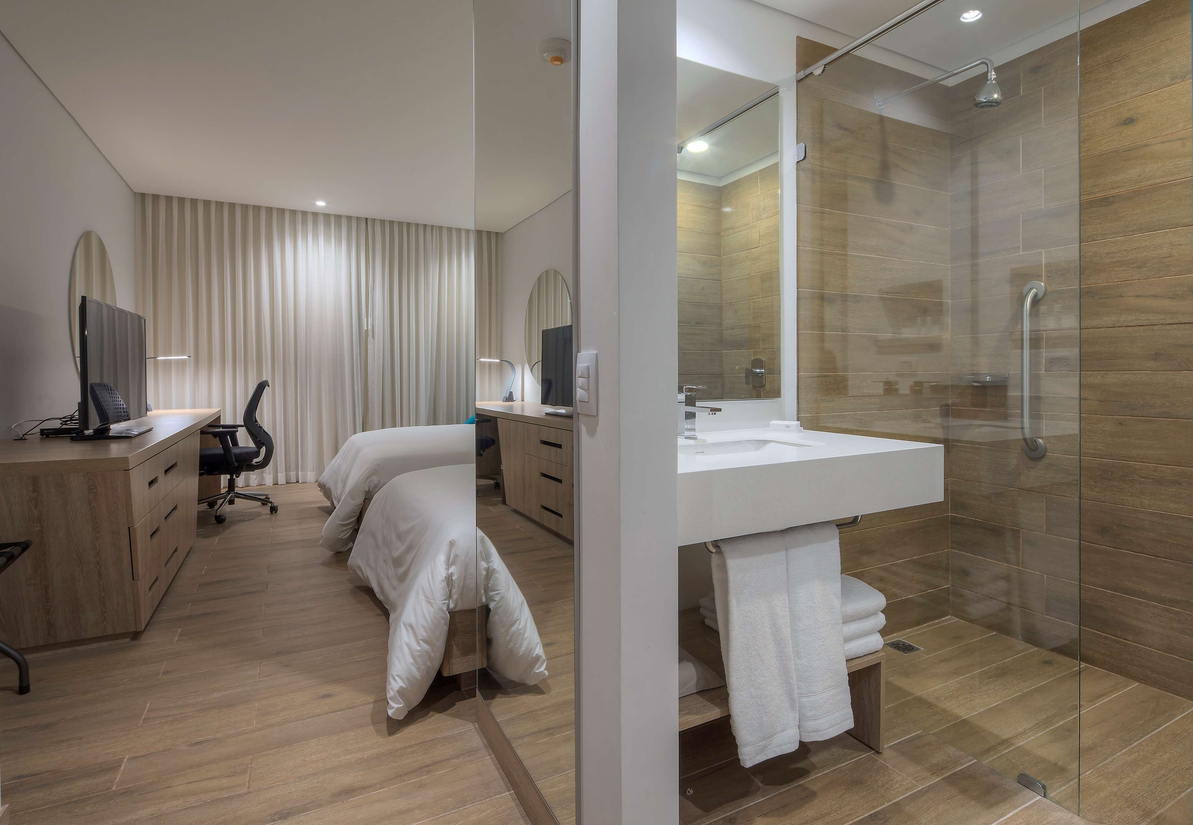 A modern hotel room with a bed, desk, chair, and a glass-enclosed shower area.