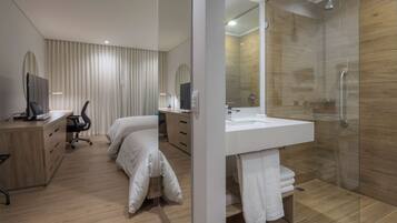 A modern hotel room with a bed, desk, chair, and a glass-enclosed shower area.