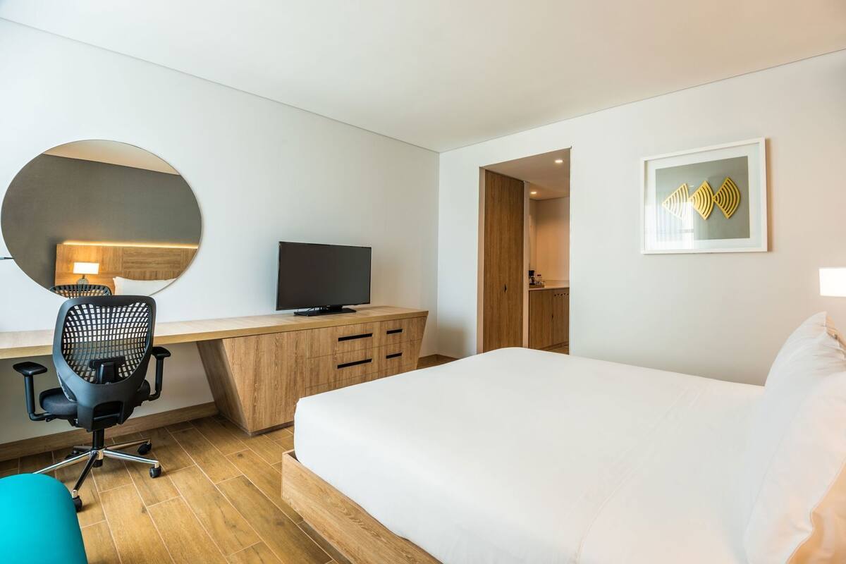A hotel room with a bed, a desk with a TV, a chair, a mirror, and a door leading to a bathroom.