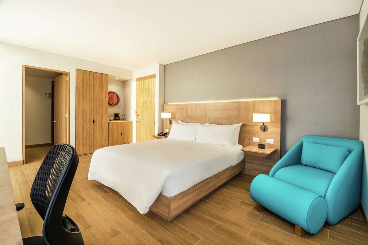 A modern hotel room with a large bed, a blue armchair, a wooden desk, and a desk fan.