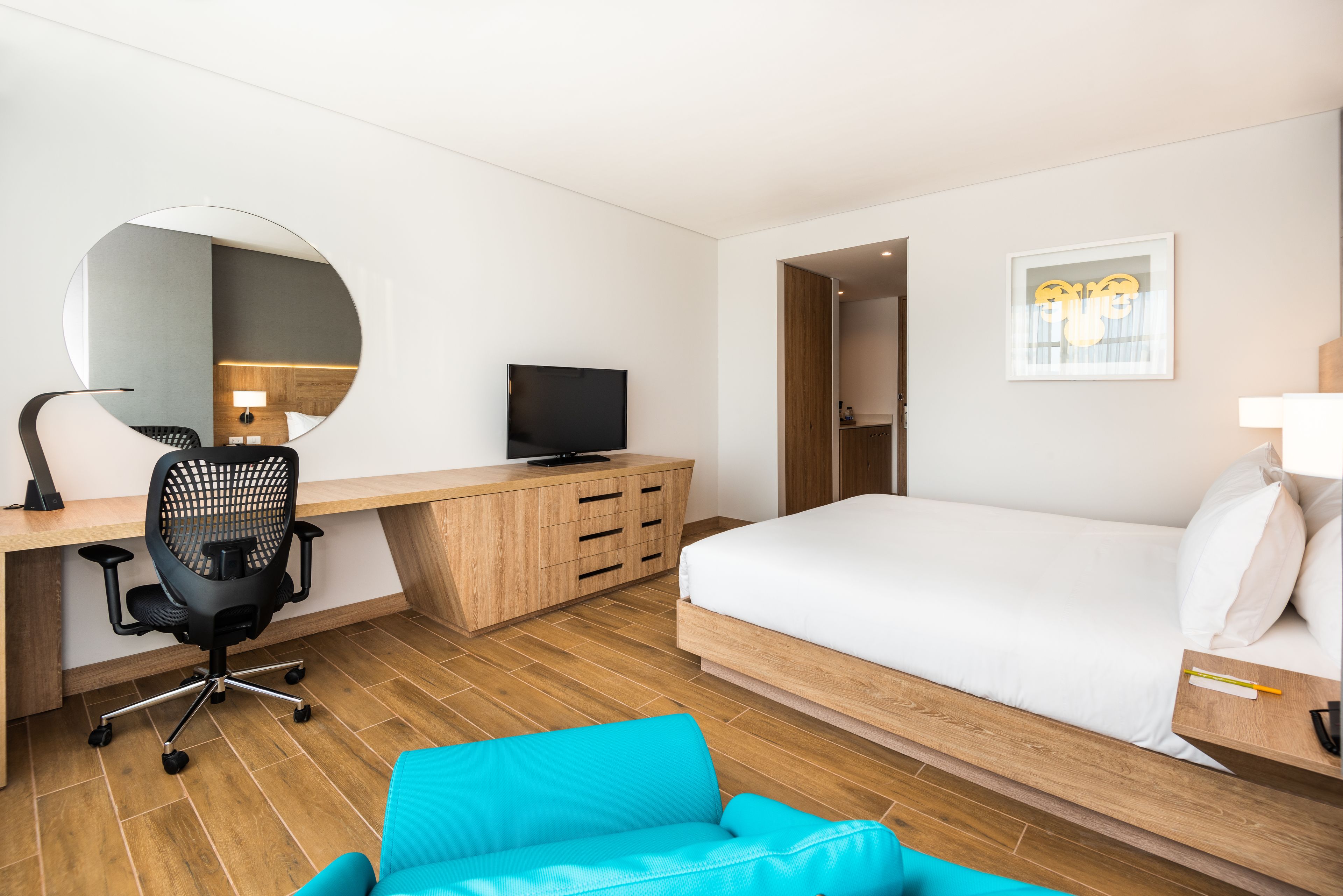 A modern hotel room with a wooden desk, a black office chair, a television, a bed with white bedding, a blue armchair, and a round mirror on the wall.
