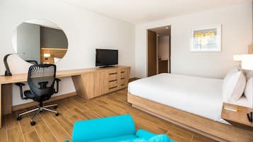 Une chambre d’hôtel moderne avec un bureau en bois, un fauteuil de bureau noir, une télévision, un lit avec une literie blanche, un fauteuil bleu et un miroir rond accroché au mur.