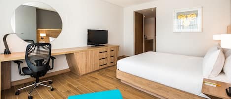 Une chambre d’hôtel moderne avec un bureau en bois, un fauteuil de bureau noir, une télévision, un lit avec une literie blanche, un fauteuil bleu et un miroir rond accroché au mur.