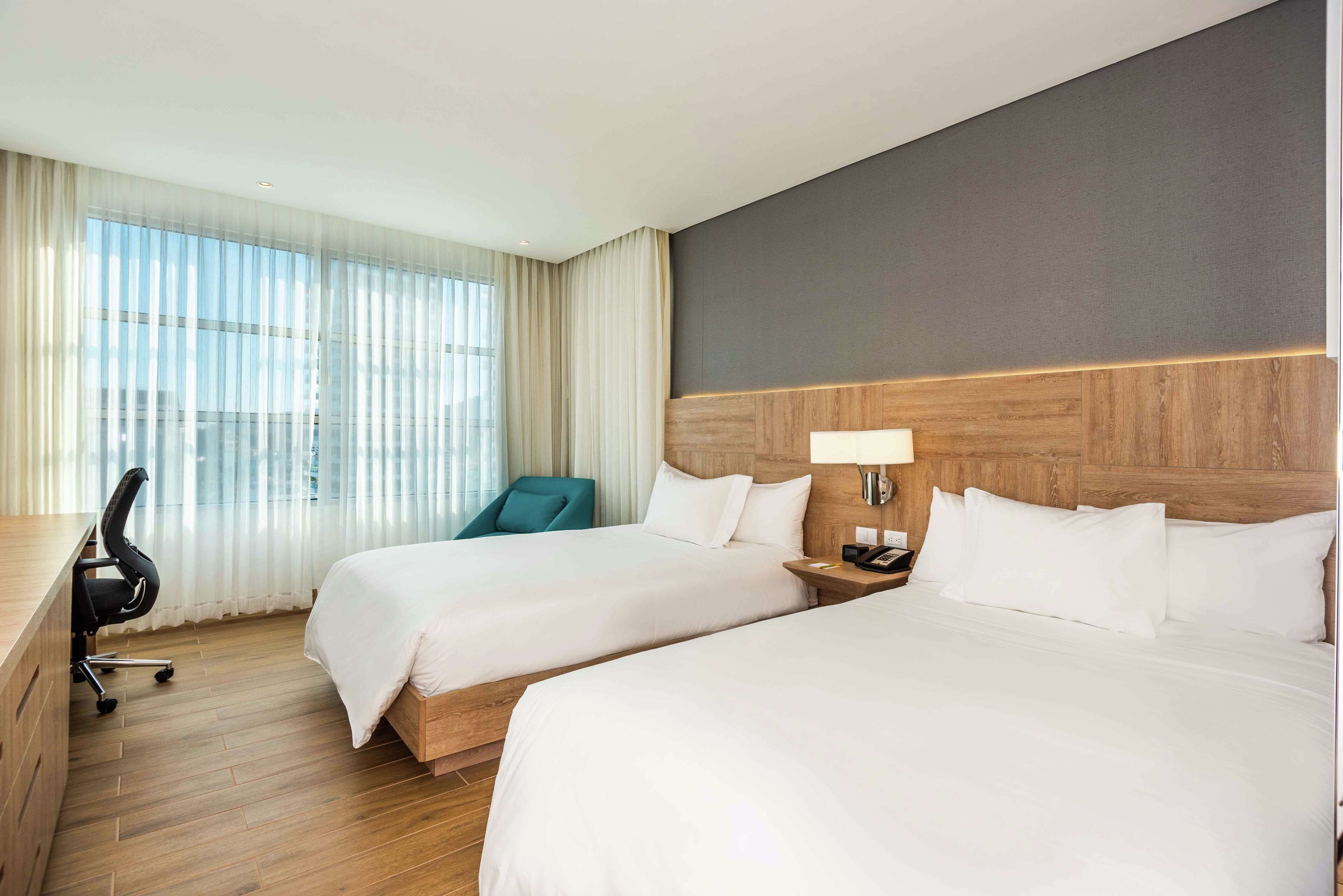 A hotel room with two beds, a desk, a chair, and a large window with sheer curtains.