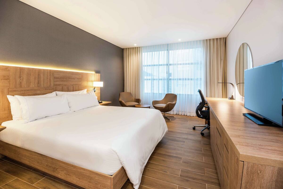 A hotel room with a large bed, a desk with a flat-screen TV, two chairs, and a large window with curtains.