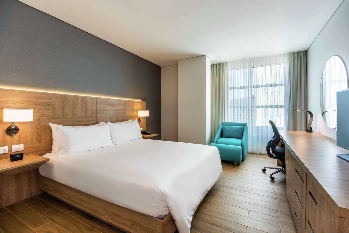 A modern hotel room with a large bed, wooden headboard, bedside lamps, a teal armchair, a desk with a chair, and a large window with curtains.